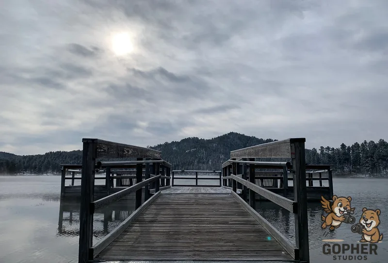 A wooden pier extends into a still lake beneath a cloudy sky and wooded hills.