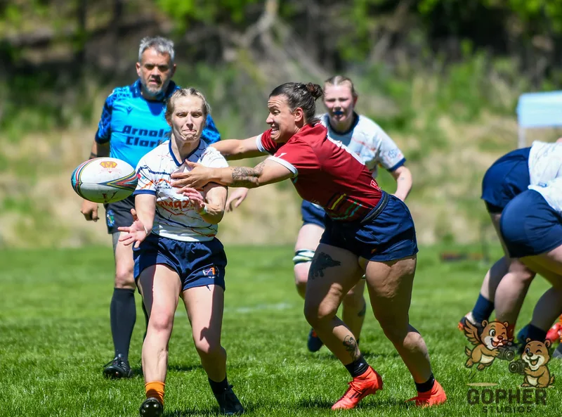 A rugby player in white passes the ball as an opponent in red reaches to tackle.